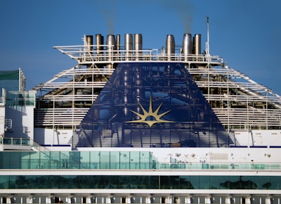 The image depicts the upper part of a cruise ship with smoke stacks and the ship's emblem prominently displayed. The structure is large and industrial, with a blue panel featuring a yellow sun-like symbol. Glass railings and other details of the ship's architecture are visible.