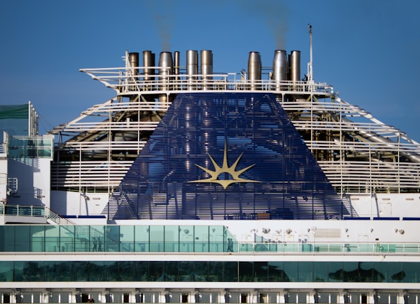 The image depicts the upper part of a cruise ship with smoke stacks and the ship's emblem prominently displayed. The structure is large and industrial, with a blue panel featuring a yellow sun-like symbol. Glass railings and other details of the ship's architecture are visible.