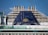 The image depicts the upper part of a cruise ship with smoke stacks and the ship's emblem prominently displayed. The structure is large and industrial, with a blue panel featuring a yellow sun-like symbol. Glass railings and other details of the ship's architecture are visible.