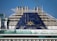 The image depicts the upper part of a cruise ship with smoke stacks and the ship's emblem prominently displayed. The structure is large and industrial, with a blue panel featuring a yellow sun-like symbol. Glass railings and other details of the ship's architecture are visible.