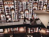 A spacious multilevel interior features large, well-lit bookshelves filled with various books and decorative items. The image captures people walking along a platform or a bridge, with some browsing the shelves. The overall lighting creates a warm and inviting atmosphere, emphasizing the vibrant colors of the items on display.