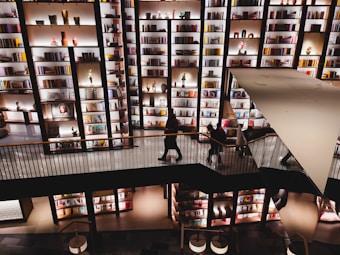 A spacious multilevel interior features large, well-lit bookshelves filled with various books and decorative items. The image captures people walking along a platform or a bridge, with some browsing the shelves. The overall lighting creates a warm and inviting atmosphere, emphasizing the vibrant colors of the items on display.