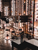 A spacious and well-lit bookstore with a modern design. Tall bookshelves filled with various colorful books cover the walls. The floor consists of neatly arranged shelves and display tables with additional lighting hanging above them. A staircase leads to a higher level surrounded by black railings.