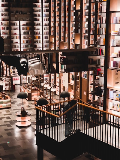 A spacious and well-lit bookstore with a modern design. Tall bookshelves filled with various colorful books cover the walls. The floor consists of neatly arranged shelves and display tables with additional lighting hanging above them. A staircase leads to a higher level surrounded by black railings.