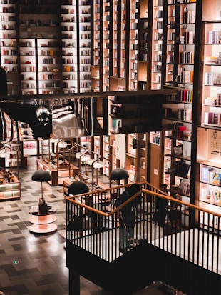 A spacious and well-lit bookstore with a modern design. Tall bookshelves filled with various colorful books cover the walls. The floor consists of neatly arranged shelves and display tables with additional lighting hanging above them. A staircase leads to a higher level surrounded by black railings.