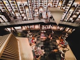 A modern bookstore with multiple levels, featuring shelves filled with books and decorative items. The shelves are illuminated with warm lighting, creating a cozy atmosphere. People are browsing or walking along the upper and lower levels.
