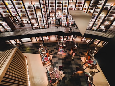 A modern bookstore with multiple levels, featuring shelves filled with books and decorative items. The shelves are illuminated with warm lighting, creating a cozy atmosphere. People are browsing or walking along the upper and lower levels.