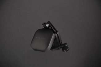 A sleek, black pair of wireless earbuds housed in a compact charging case, positioned against a dark background. The earbuds have a minimalist design with smooth curves, and the brand name 'Haylou' is faintly visible on the case.