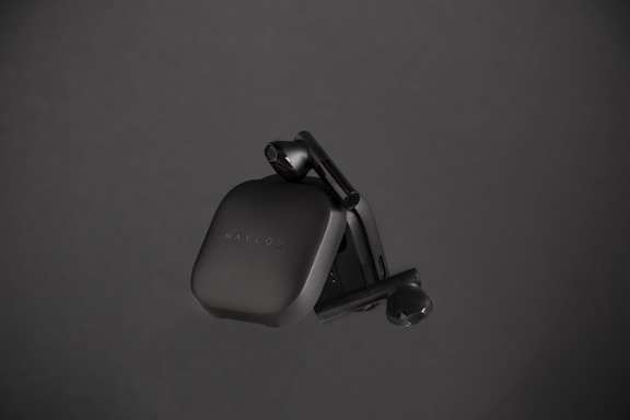 A sleek, black pair of wireless earbuds housed in a compact charging case, positioned against a dark background. The earbuds have a minimalist design with smooth curves, and the brand name 'Haylou' is faintly visible on the case.