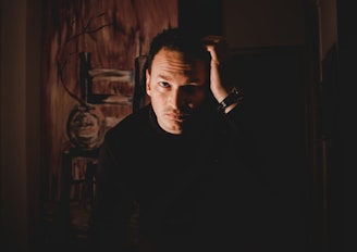 A person in a dark setting with a contemplative expression, hand placed on their head. The background features abstract art with brown and earthy tones.