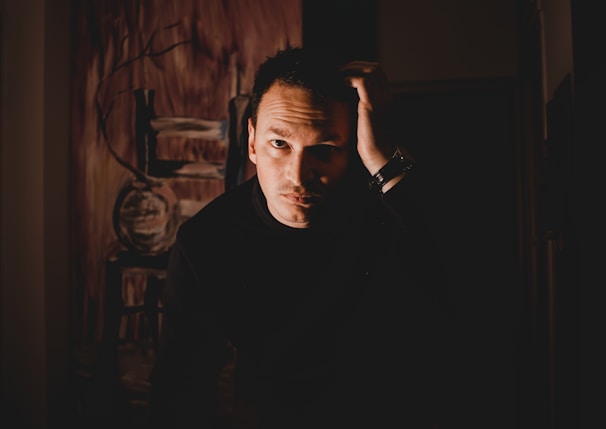 A person in a dark setting with a contemplative expression, hand placed on their head. The background features abstract art with brown and earthy tones.