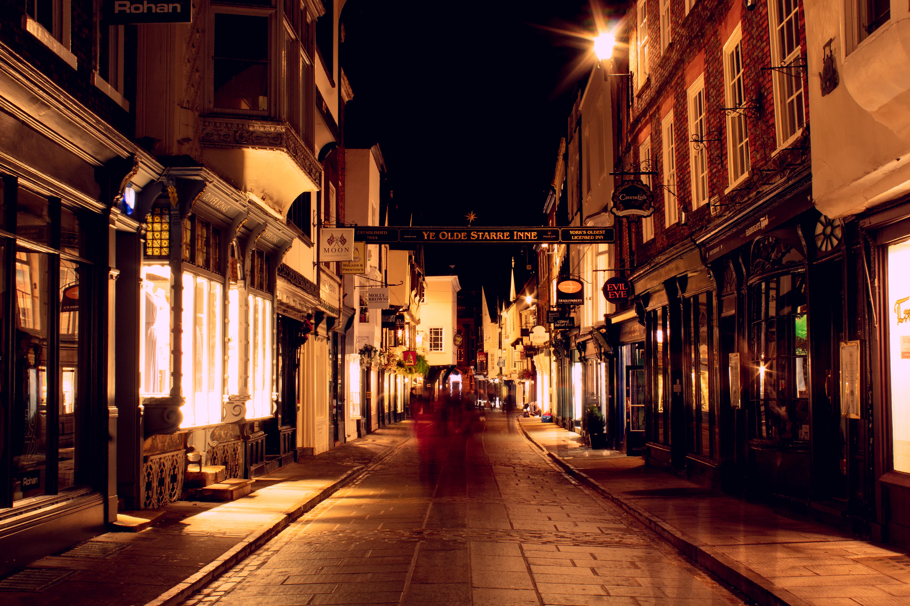 Stonegate in York at night, its cobbles and timber-framed shopfronts washed in warm lamplight beneath the hanging gallows sign of Ye Olde Starre Inne.