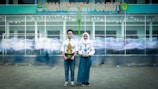 A young man and woman in school uniforms stand holding trophies in front of a school building with the sign 'SMA NEGERI 19 GARUT.' In the background, blurred figures of other students in motion create a dynamic sense of movement.