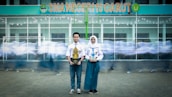 A young man and woman in school uniforms stand holding trophies in front of a school building with the sign 'SMA NEGERI 19 GARUT.' In the background, blurred figures of other students in motion create a dynamic sense of movement.
