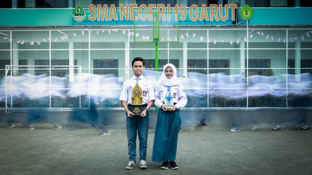 A young man and woman in school uniforms stand holding trophies in front of a school building with the sign 'SMA NEGERI 19 GARUT.' In the background, blurred figures of other students in motion create a dynamic sense of movement.