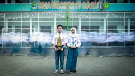 A young man and woman in school uniforms stand holding trophies in front of a school building with the sign 'SMA NEGERI 19 GARUT.' In the background, blurred figures of other students in motion create a dynamic sense of movement.