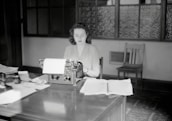 A woman sits at a mid-20th-century office desk, working on a typewriter. The room has a vintage feel with wooden furniture and frosted glass windows. Papers are spread out on the desk, and there are various office supplies.
