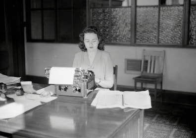 A woman sits at a mid-20th-century office desk, working on a typewriter. The room has a vintage feel with wooden furniture and frosted glass windows. Papers are spread out on the desk, and there are various office supplies.