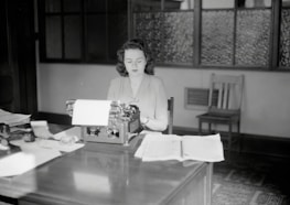 A woman sits at a mid-20th-century office desk, working on a typewriter. The room has a vintage feel with wooden furniture and frosted glass windows. Papers are spread out on the desk, and there are various office supplies.
