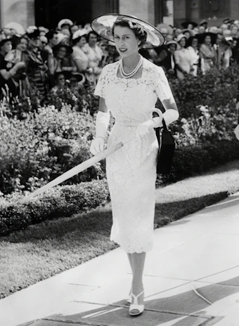 A fashionable woman in a flowing white formal dress with delicate lace details, walking through a sunlit European garden.