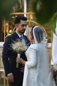 A bride and groom are standing closely together. The groom is wearing a dark suit and tie, and the bride is in a white gown with a sparkling veil. The bride holds a bouquet of dried flowers. The background features a decorative structure with flowers.