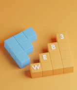 A three-dimensional arrangement of blocks on a solid orange background. The blue blocks are shaped like an arrow pointing left, and the orange blocks are stacked like steps with white letters spelling 'WEB 3'.