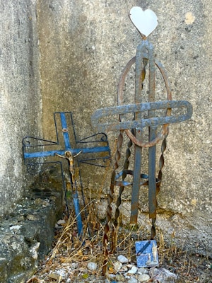 Two intricate metal crosses are situated against a weathered stone wall. One cross has a stylized depiction of Jesus, while the other features a heart-shaped ornament at the top. The ground is scattered with dry leaves and small stones, lending an aged, rustic charm to the scene.