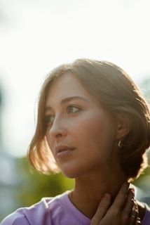 A contemplative individual with short, light brown hair gazes thoughtfully into the distance. Sunlight softly illuminates the side of the face, creating a warm and serene atmosphere. The person is wearing a light-colored top and gently holds their hand near their neck.
