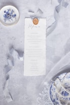 A decorative menu card is styled with a wax seal and elegant ribbon, placed on a textured gray surface. Accompanying the menu is a small dish with blue floral designs, containing hairpins with pearls, nearby a second dish decorated similarly is filled with pearls. Delicate lace fabric stretches across the surface.