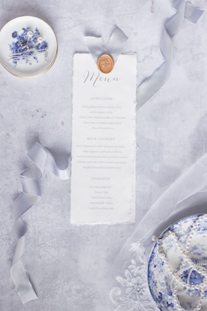 A decorative menu card is styled with a wax seal and elegant ribbon, placed on a textured gray surface. Accompanying the menu is a small dish with blue floral designs, containing hairpins with pearls, nearby a second dish decorated similarly is filled with pearls. Delicate lace fabric stretches across the surface.