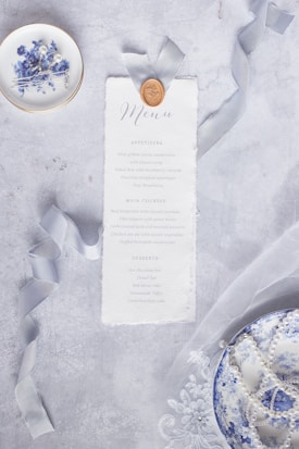 A decorative menu card is styled with a wax seal and elegant ribbon, placed on a textured gray surface. Accompanying the menu is a small dish with blue floral designs, containing hairpins with pearls, nearby a second dish decorated similarly is filled with pearls. Delicate lace fabric stretches across the surface.