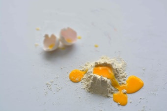 A cracked egg is open with the yolk and some of the egg white spreading on a flat surface. Nearby, there are two empty eggshell halves. A small mound of flour surrounds the egg yolk and is scattered on the surface.