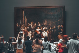 A group of people are gathered in front of a large, framed painting in a museum. The painting appears to be a historical or classical artwork, with numerous figures depicted. Visitors are observing and taking photos with their phones, showing interest and engagement with the art piece.