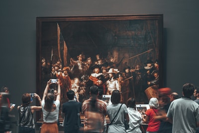 A group of people are gathered in front of a large, framed painting in a museum. The painting appears to be a historical or classical artwork, with numerous figures depicted. Visitors are observing and taking photos with their phones, showing interest and engagement with the art piece.