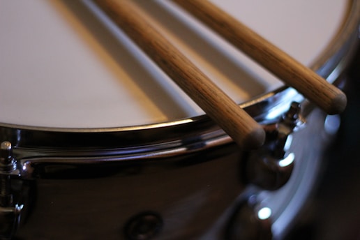 Two wooden drumsticks are resting on the surface of a shiny snare drum. The metal rim of the drum reflects light, adding to the instrument's polished appearance.