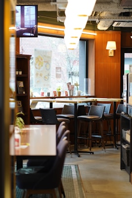 A cozy café interior with wooden paneling and large windows letting in natural light. The space features high tables with chairs, wall decor, and neatly arranged items on the counters. A television screen is mounted near the ceiling displaying a serene scene. Warm lighting fixtures enhance the inviting atmosphere.