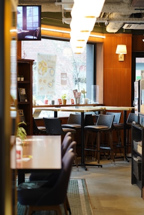 A cozy café interior with wooden paneling and large windows letting in natural light. The space features high tables with chairs, wall decor, and neatly arranged items on the counters. A television screen is mounted near the ceiling displaying a serene scene. Warm lighting fixtures enhance the inviting atmosphere.
