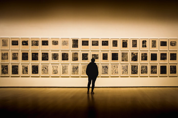 A person stands in a silent, well-lit art gallery observing a large collection of framed artworks arranged in a grid on a light-colored wall. The artworks appear to be abstract prints, each with distinct patterns and textures. The room has a wooden floor, and the overall atmosphere is contemplative.