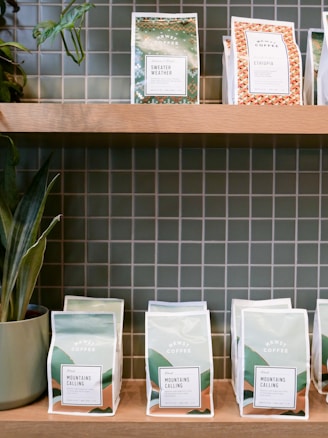 Several bags of coffee displayed on wooden shelves against a tiled wall backdrop. The bags feature different designs and labels, including 'Sweater Weather' and 'Ethiopia'. There are also potted plants adding a touch of greenery.