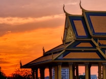 A traditional Southeast Asian temple structure with a tiered roof and ornate architectural details stands against a dramatic sunset sky. The silhouette of the building is highlighted by the warm orange and red hues of the sunset, creating a striking contrast.