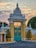 An ornate, architectural gate with elaborate decorations and intricate designs stands prominently. The gate is flanked by manicured, symmetrical bushes in golden planters, set against a backdrop of a softly hued sky at dawn or dusk. Detailed metalwork features on the closed turquoise gates, highlighted by circular, gold accents.