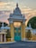 An ornate, architectural gate with elaborate decorations and intricate designs stands prominently. The gate is flanked by manicured, symmetrical bushes in golden planters, set against a backdrop of a softly hued sky at dawn or dusk. Detailed metalwork features on the closed turquoise gates, highlighted by circular, gold accents.