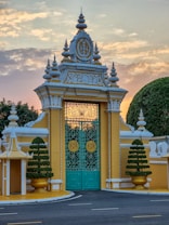 An ornate, architectural gate with elaborate decorations and intricate designs stands prominently. The gate is flanked by manicured, symmetrical bushes in golden planters, set against a backdrop of a softly hued sky at dawn or dusk. Detailed metalwork features on the closed turquoise gates, highlighted by circular, gold accents.