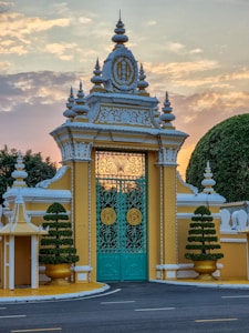 An ornate, architectural gate with elaborate decorations and intricate designs stands prominently. The gate is flanked by manicured, symmetrical bushes in golden planters, set against a backdrop of a softly hued sky at dawn or dusk. Detailed metalwork features on the closed turquoise gates, highlighted by circular, gold accents.