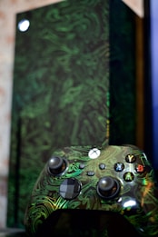 A close-up of a video game controller with a unique green swirling pattern and buttons labeled with letters. In the background is a matching console with a similar design, partially blurred.