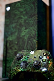 A close-up of a video game controller with a unique green swirling pattern and buttons labeled with letters. In the background is a matching console with a similar design, partially blurred.