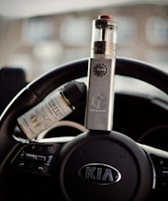 A vape device is placed upright on a car's steering wheel, with a bottle of e-liquid resting beside it. The steering wheel belongs to a Kia vehicle, as indicated by the emblem in the center.