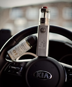A vape device is placed upright on a car's steering wheel, with a bottle of e-liquid resting beside it. The steering wheel belongs to a Kia vehicle, as indicated by the emblem in the center.