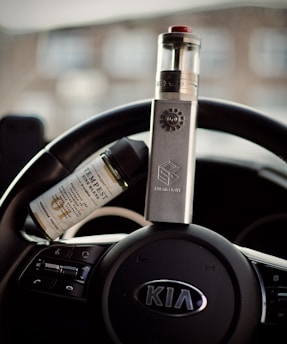 A vape device is placed upright on a car's steering wheel, with a bottle of e-liquid resting beside it. The steering wheel belongs to a Kia vehicle, as indicated by the emblem in the center.