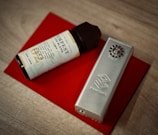 A cylindrical vape juice bottle with a light-colored label and a black cap is placed next to a metallic vape mod. Both items are resting on a red rectangular surface that contrasts with the wooden tabletop underneath.